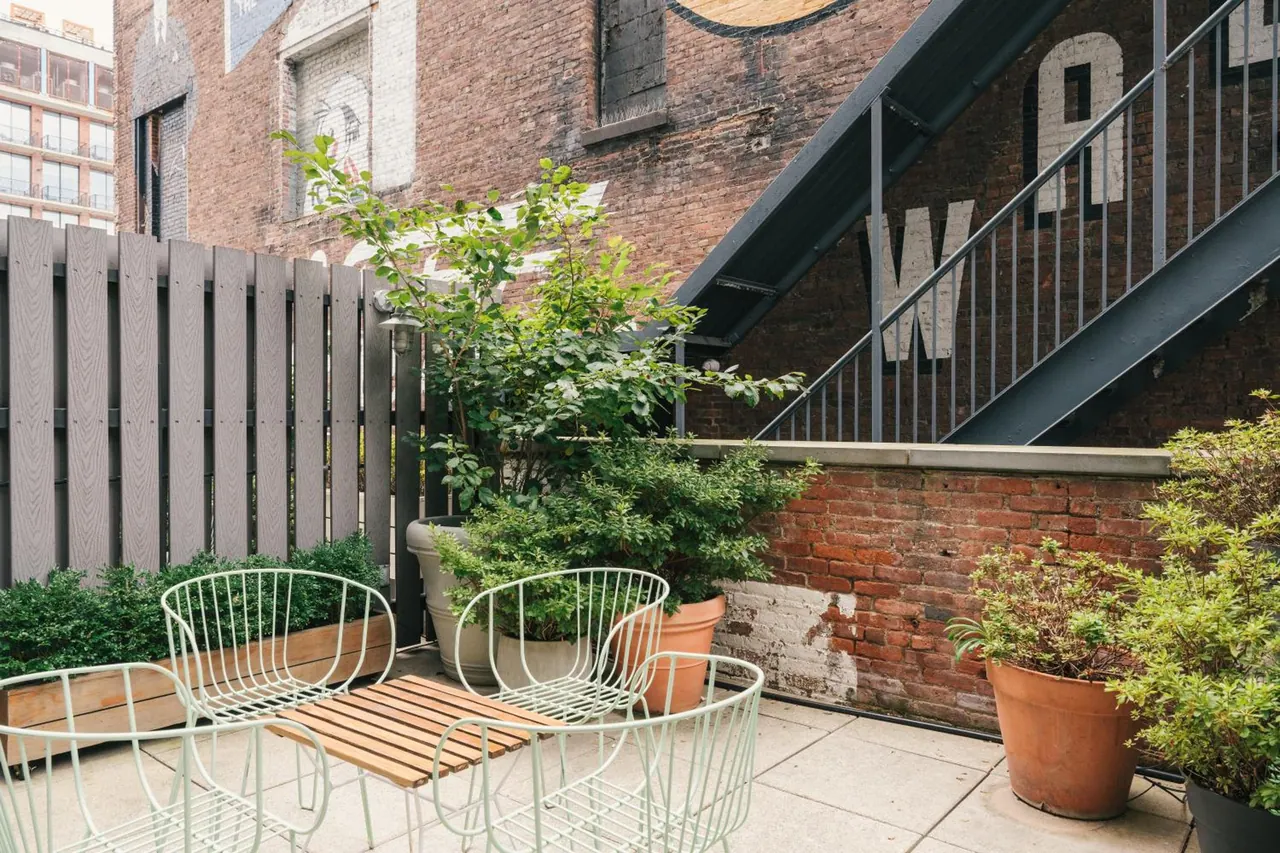 A furnished outdoor terrace with mint green wire chairs, a wooden table, potted plants, a wooden fence and a brick wall with a fire escape.
