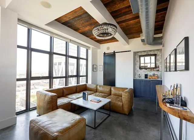 A living area with a brown leather sectional sofa, a marble coffee table, a wood-panelled ceiling and floor-to-ceiling windows looking out over the city.