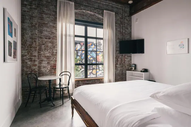 A bedroom with a made bed, exposed brick wall, round bistro table with two black chairs, wall-mounted television, and a window looking onto a mural-covered building.