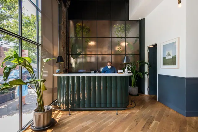 A green fluted reception desk stands in a lobby with floor-to-ceiling windows, and a masked staff member sits behind the counter.