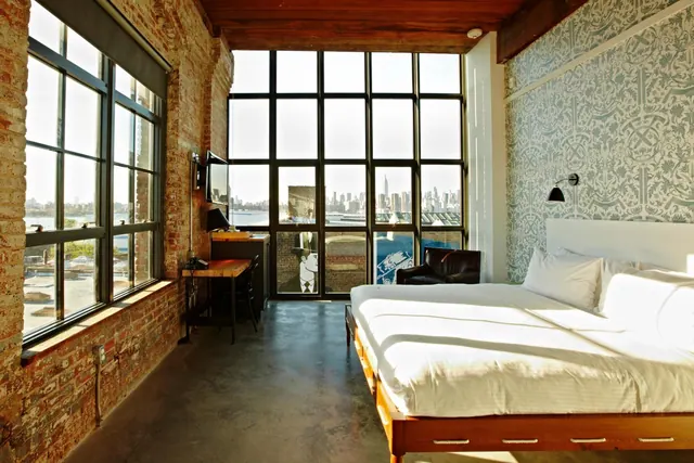A bedroom with a king bed, exposed brick walls, a work desk and a large window wall looking across the water toward the Manhattan skyline.