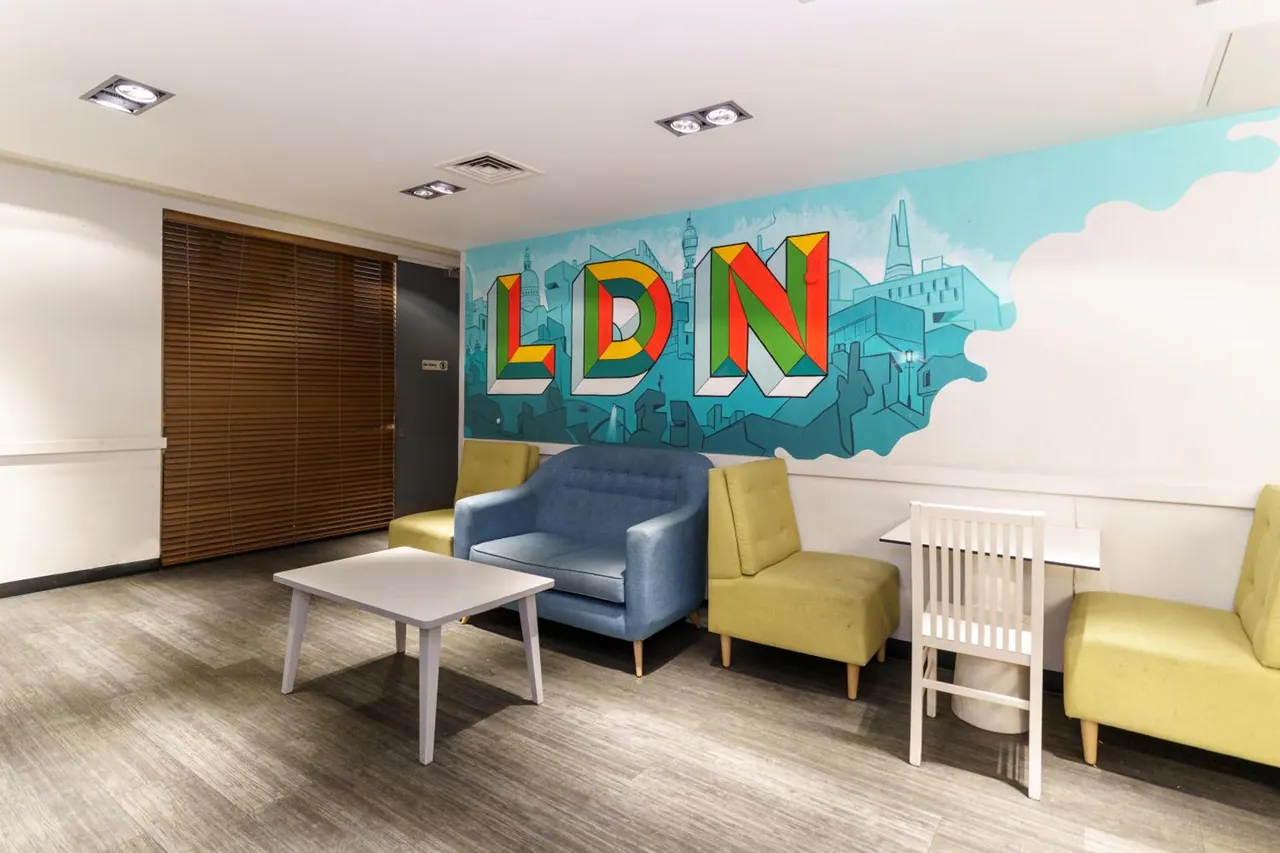 A lounge seating area with yellow and blue armchairs, white tables and a wall mural with the letters LDN and a painted city skyline.