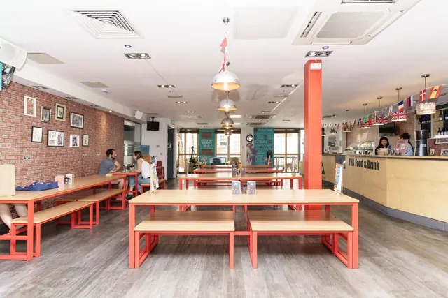 A large communal dining room with long red-framed wooden tables and benches, a brick wall of framed pictures, and a food and drink counter on the right with flags hanging above it.