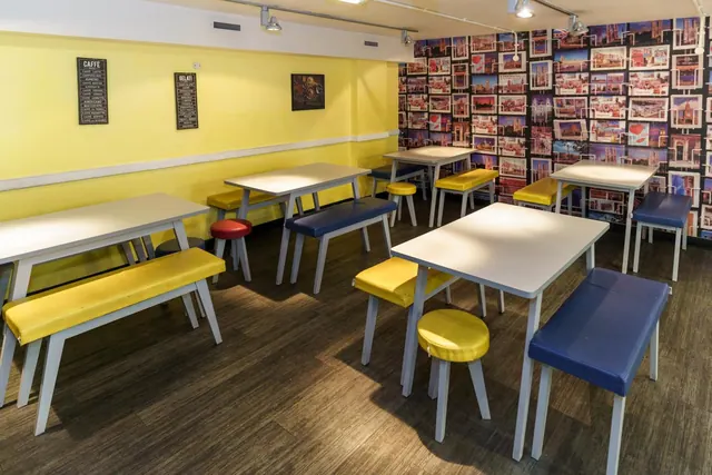 A dining room with yellow walls, white tables, yellow and blue benches and stools, and a wall covered in framed postcards.