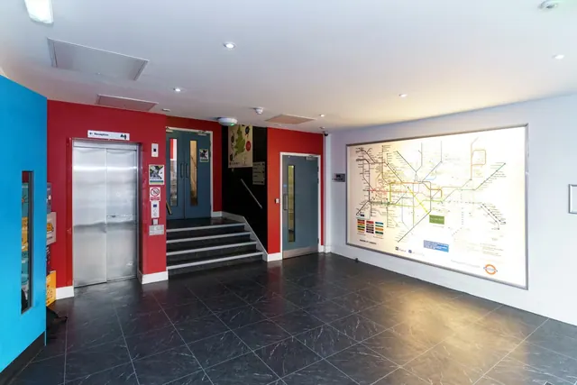 A lobby with red walls, metal lift doors, a staircase with dark tiled steps, and a large backlit London Underground map on the right wall.