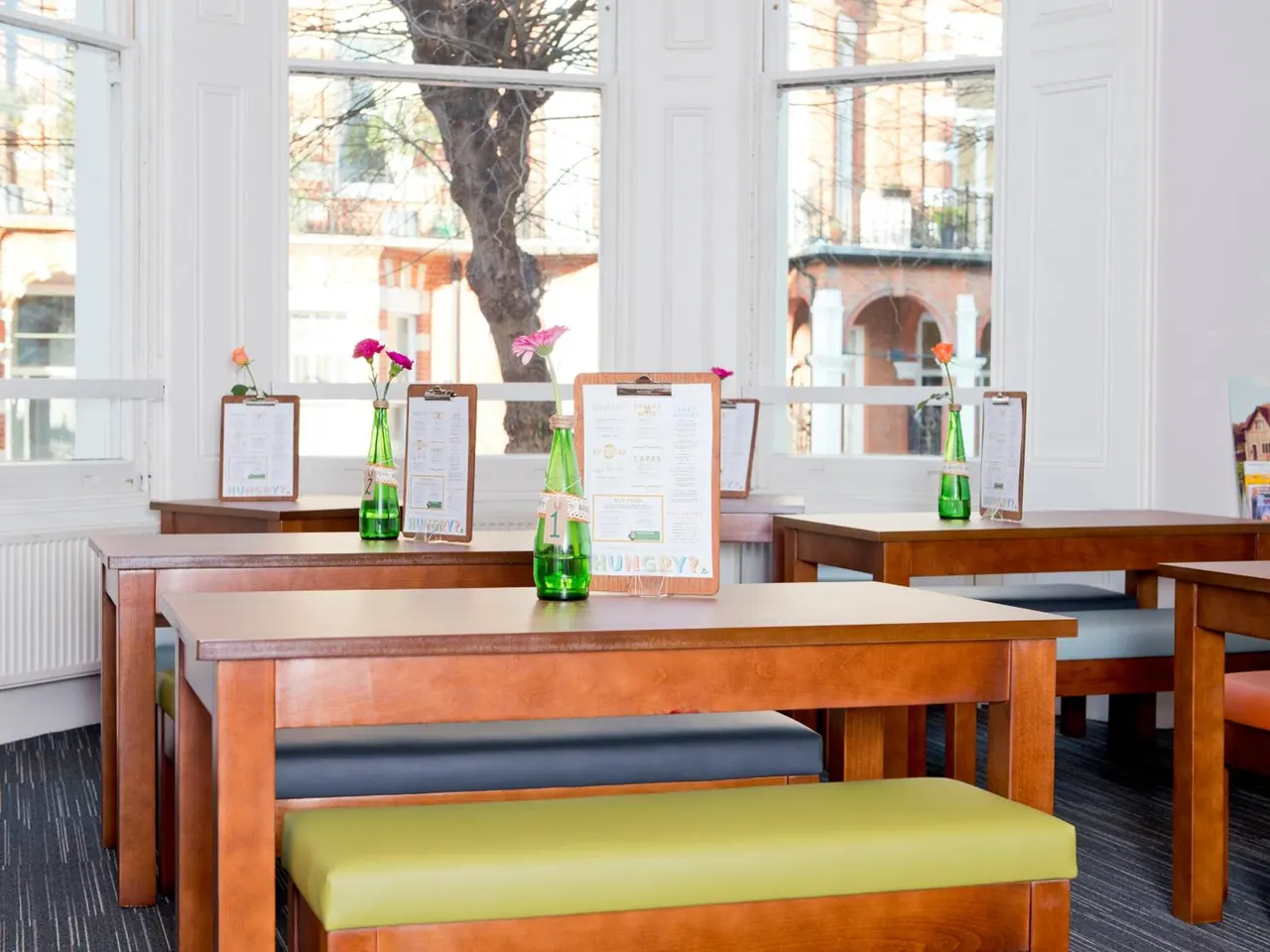 An empty dining room with wooden tables, green and grey bench seating, menus on clipboards, and green bottles with flowers in front of tall windows.