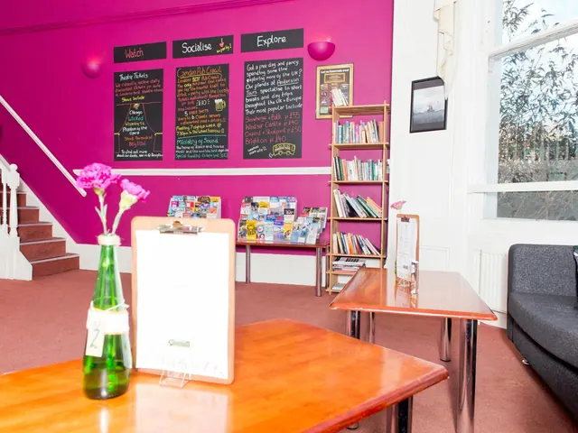 A lounge area with a magenta wall, chalkboard signs, a bookshelf, a brochure rack, wooden tables and a grey sofa.