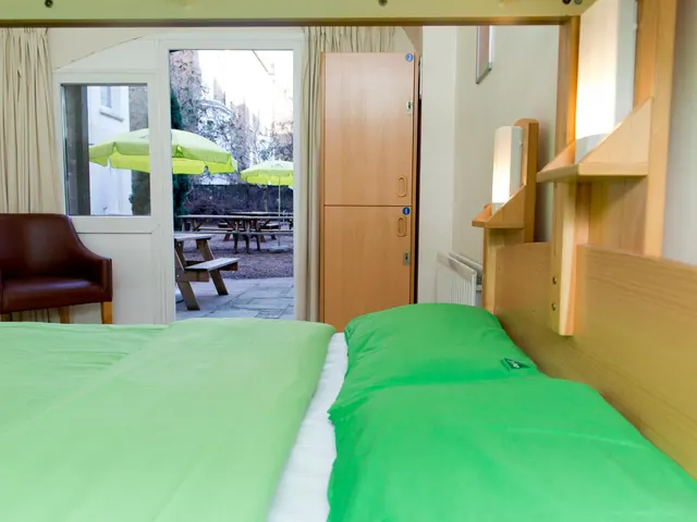 A bedroom with green bedding and a wooden headboard, with a leather armchair, wooden lockers, and a glass door onto a courtyard with picnic tables and green umbrellas.