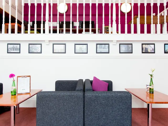A lounge area with grey sofas, a purple cushion, wooden side tables with flower vases, framed prints on the wall and a white balcony railing above.