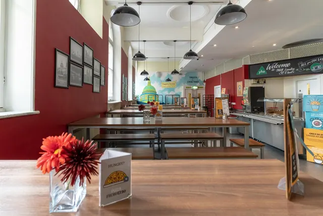 A communal dining room with long wooden tables and benches, pendant lights, and a serving counter below a Welcome sign for YHA London St Paul's.