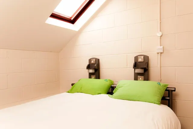 A double bed with white bedding and two green pillows sits under a sloped ceiling with a skylight, beside wall-mounted shelves with sockets.