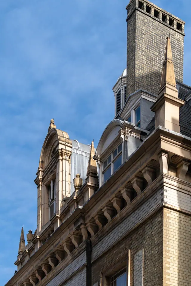 The upper facade and roofline of a brick and stone building, with dormer windows, a chimney stack and an ornate cornice, against a blue sky.