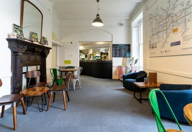 A lounge area with grey carpet, blue armchairs, wooden tables and mismatched chairs, a tube map on the wall, and a bar counter in the background.