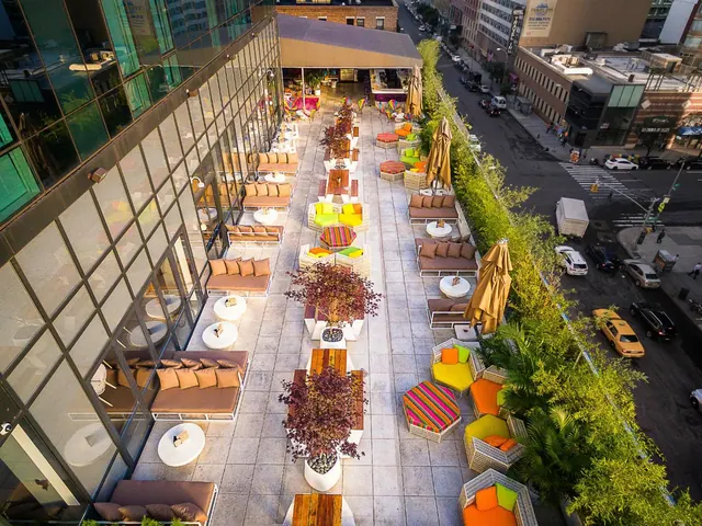 Aerial view of an outdoor terrace with sofas, colourful cushioned seating, wooden tables and folded umbrellas, with a street and cars visible below.