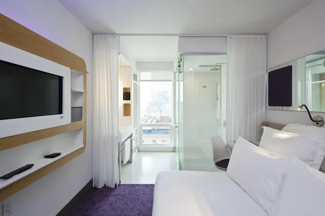 A bedroom with a made bed, a wall-mounted television, a glass-enclosed shower and a window showing city buildings.