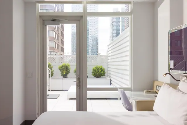 A bedroom with a white bed in the foreground, a glass door and windows opening onto a private terrace with planters, and city towers visible beyond.