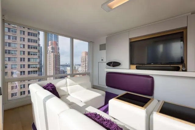 A living area with white leather sofas, purple cushions, a wall-mounted flat-screen television and floor-to-ceiling windows overlooking city high-rise buildings.