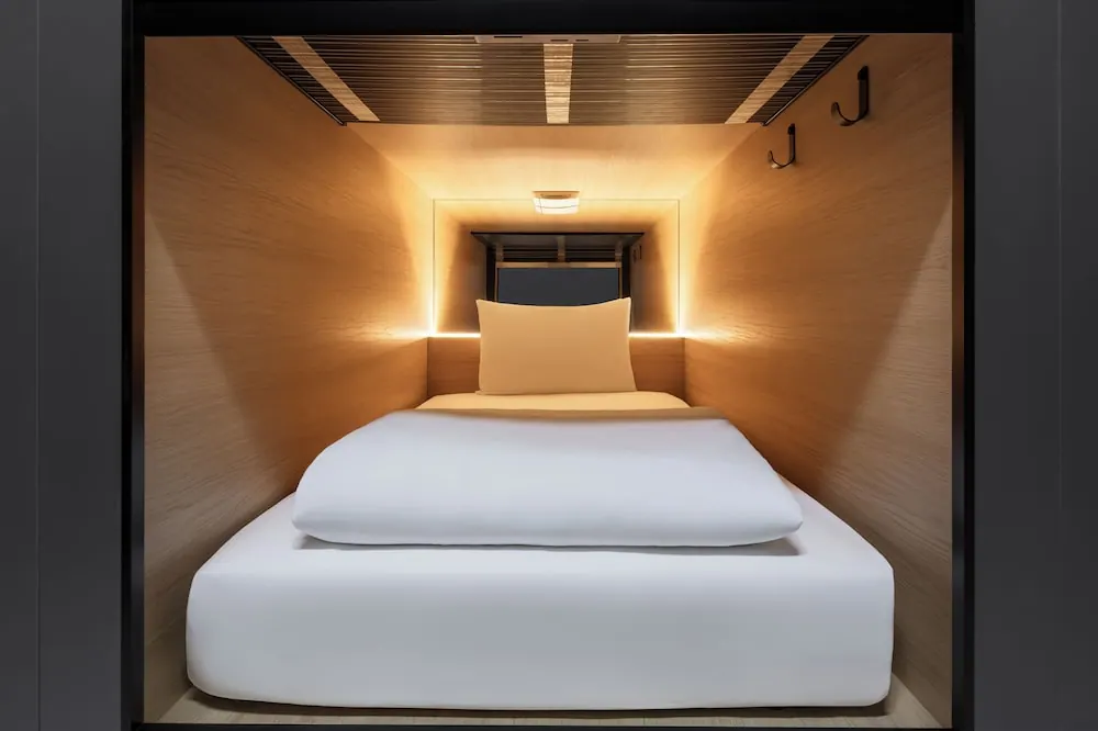 A single bed with white linen and an orange pillow sits inside a wood-lined sleeping pod with warm built-in lighting and wall hooks.