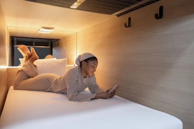 A woman lies on a white bed in a wood-panelled sleeping pod, looking at a tablet.