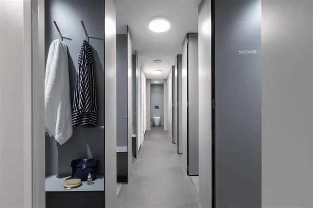 A narrow grey corridor with cubicle doors, a door labelled SHOWER, a towel and robe on wall hooks, and a toilet visible at the far end.