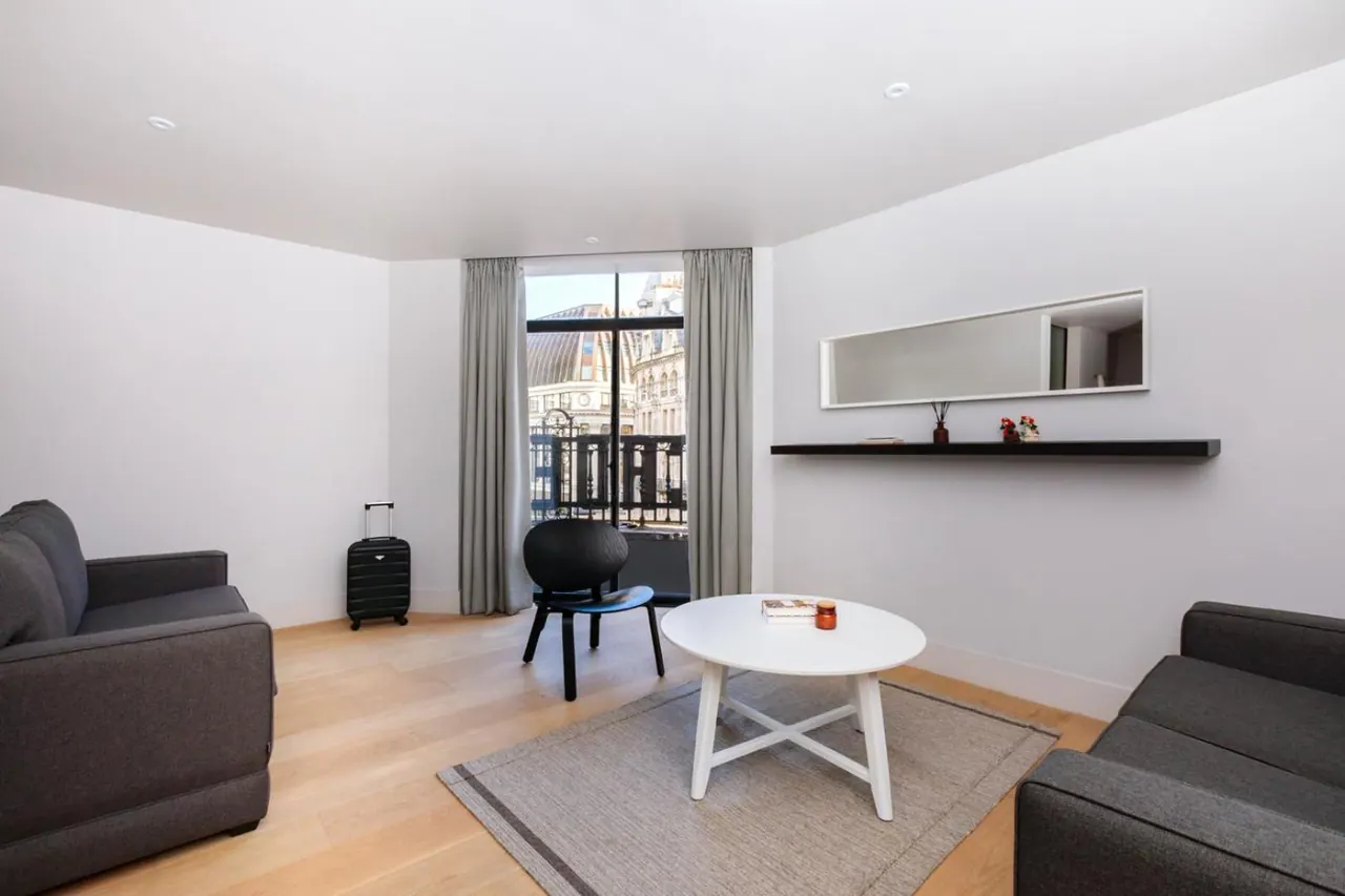 A living area with two grey sofas, a white round coffee table, a black chair and a black suitcase, with a tall window looking out onto a building with a domed roof.