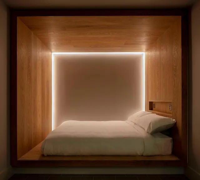 A made bed sits on a raised wooden platform inside a wood-panelled alcove, with a white LED strip outlining the recessed wall behind it.