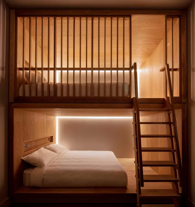 A bedroom with a timber bunk arrangement: a double bed on a platform below and an upper sleeping platform behind slatted railings, lit by warm LED strips and reached by a wooden ladder.