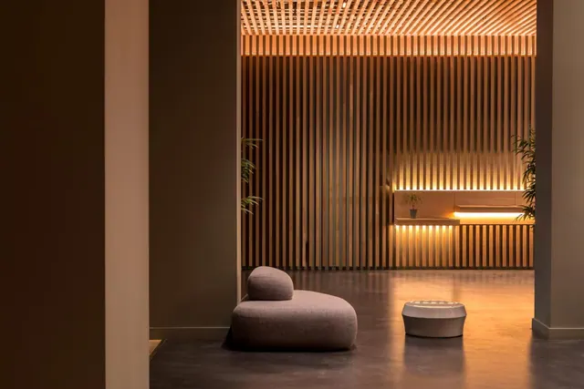 A grey upholstered lounge seat and a white round stool sit on a dark floor in front of a wooden slatted wall lit from above and below.