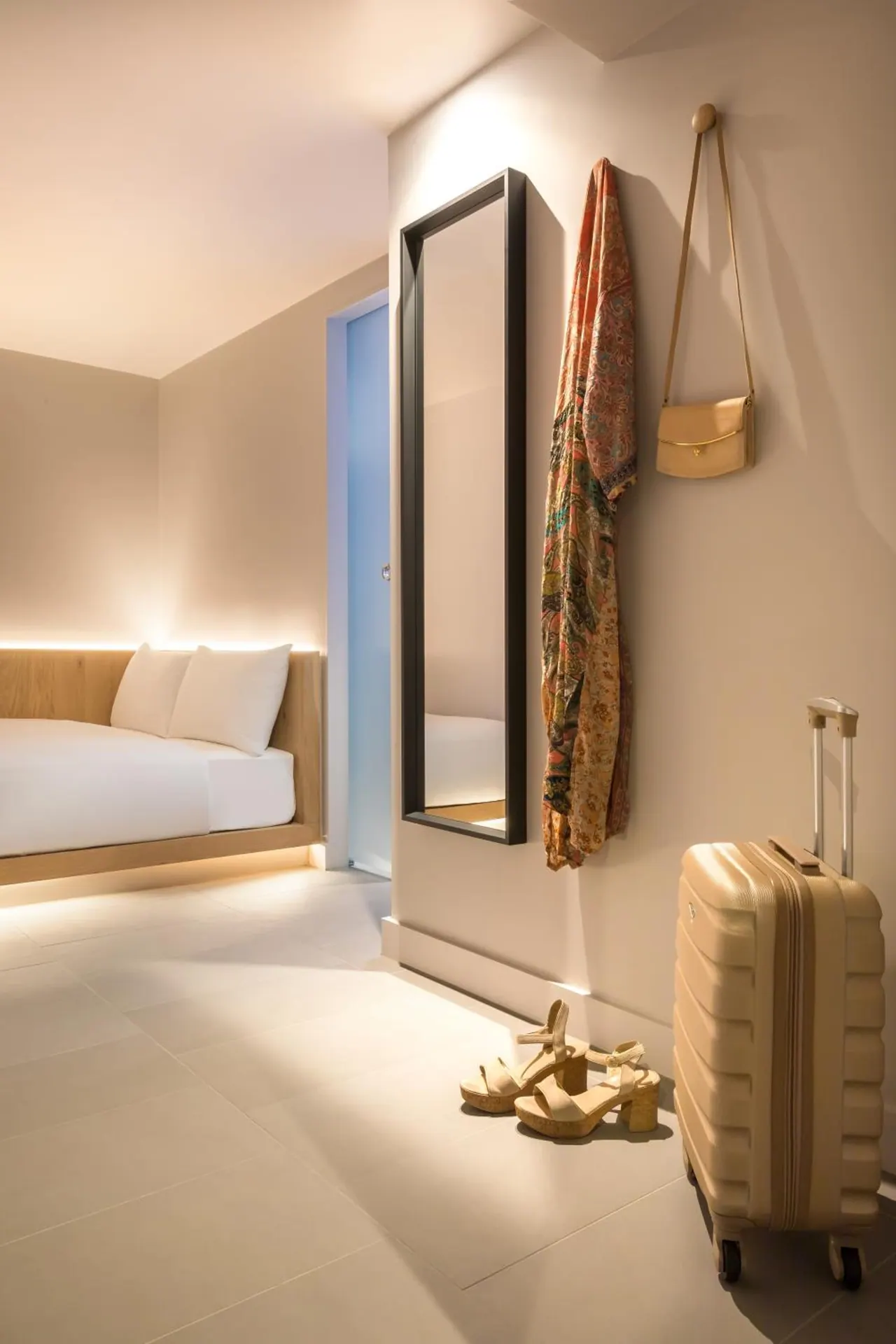 A hotel bedroom with a double bed beside a full-length mirror, a hanging scarf and bag on the wall, and a suitcase and sandals on the floor.