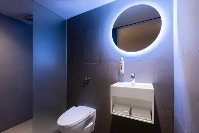 A bathroom with a wall-mounted toilet, a basin with folded towels in a wall niche, a backlit round mirror and a frosted glass shower screen.