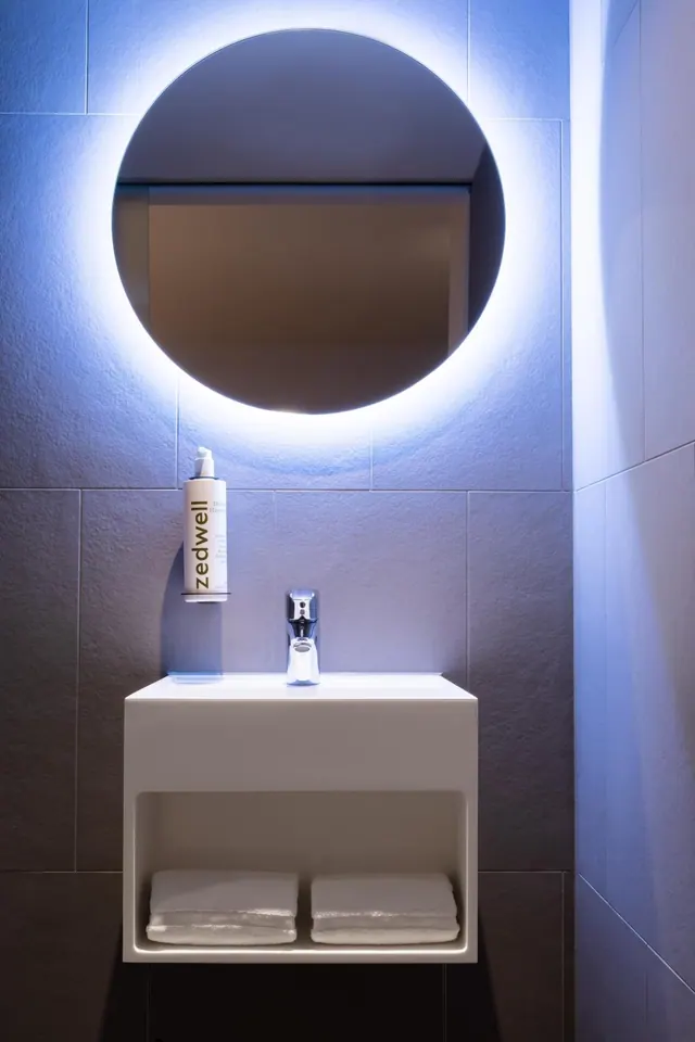 A bathroom with a round backlit mirror above a wall-mounted white basin, a zedwell bottle on the tiled wall, and two folded towels on an open shelf beneath the basin.