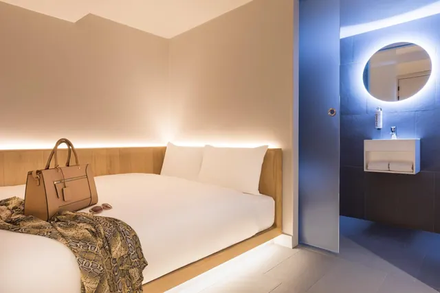 A bedroom with a raised double bed with white linen, a tan handbag and patterned throw on it, beside a bathroom with an illuminated round mirror and wall-mounted basin.