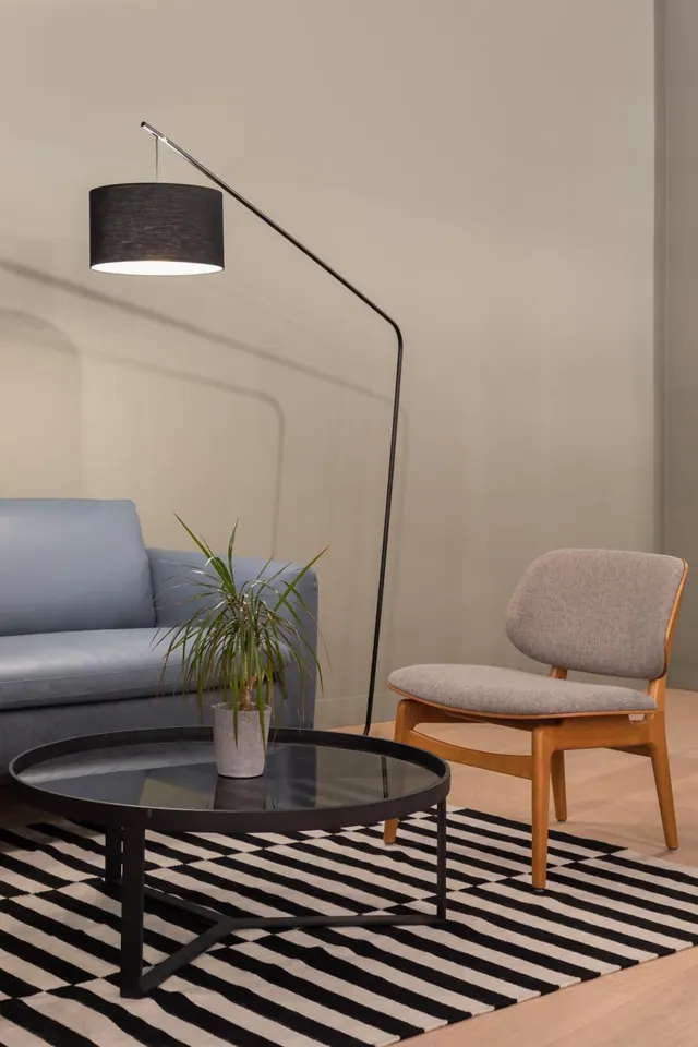 A staged living room with a blue sofa, a wooden armchair, an arc floor lamp, a potted plant, a black coffee table and a striped rug.