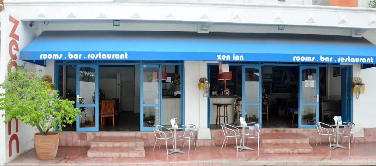 The front of the Zen Inn building shows a blue awning reading rooms, bar, restaurant, with outdoor tables and chairs and a potted plant on the terrace.