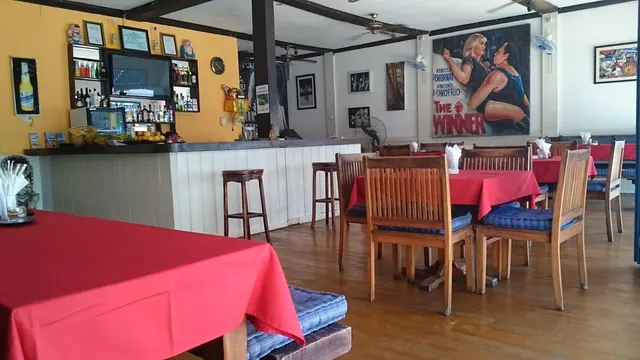 A restaurant dining room with red-clothed tables, wooden chairs with blue cushions, a bar with bottles and a television, and a large mural poster on the wall.