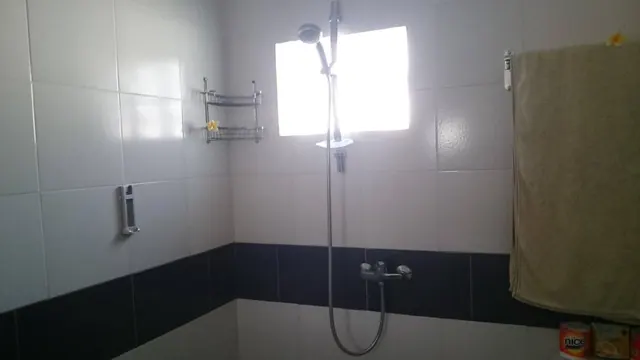 A tiled bathroom with a handheld shower on a wall bar, a small bright window, a hanging beige towel on the right, and a packaged biscuit box in the lower right corner.