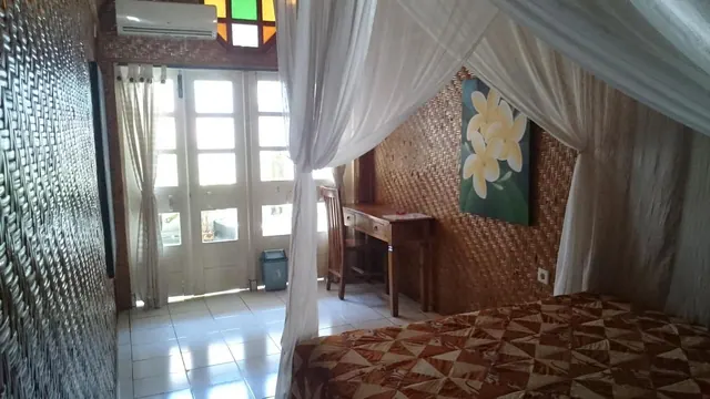 A bedroom with a bed under white canopy netting, a wooden desk and chair, woven bamboo walls, glass doors to the outside, and a wall-mounted air conditioner.