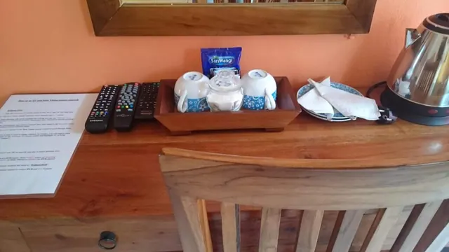 A wooden sideboard holds a tea tray with white cups, a glass sugar jar, a coffee sachet, folded napkins and an electric kettle, with remote controls and an instruction sheet on the left.