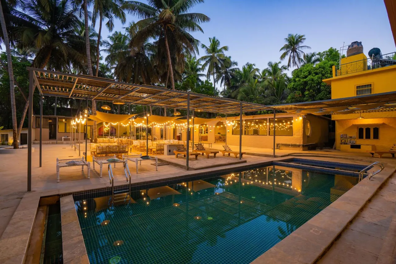 A large swimming pool sits beside a covered outdoor seating area surrounded by coconut palms.